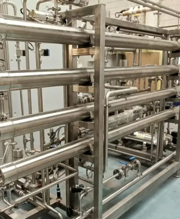 A stainless steel industrial piping and filtration system mounted on a metal frame structure inside a processing facility. The system consists of multiple horizontal pipes arranged in parallel tiers across five or six levels, supported by vertical stainless steel uprights. Various sanitary fittings, clamps, valves, and connectors join the pipes at multiple points. A cylindrical inline filter or membrane housing is visible on the right side of the lower section. The lower portion of the system features additional piping with manual valves, pressure gauges, and flow control components. The floor beneath is a clean blue-gray epoxy or tiled surface. Additional piping and equipment can be seen in the background, along with a wooden panel or wall section. The polished, sanitary-grade stainless steel construction suggests this is part of a pharmaceutical, dairy, beverage, or food processing operation, possibly a pasteurization, filtration, or heat exchange system