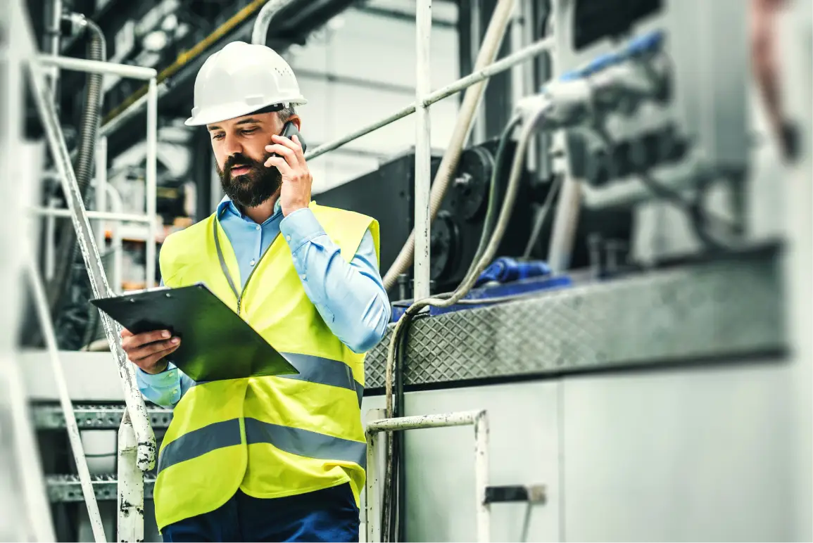 A male industrial engineer or site supervisor walking through a factory or processing plant while multitasking. He is wearing a white hard hat, a high-visibility yellow-green safety vest with reflective silver stripes, and a light blue dress shirt underneath. He has a dark beard and is holding a mobile phone to his right ear while carrying a clipboard or folder in his left hand, looking down at it as he walks. Behind him, a complex array of industrial equipment is visible, including metal pipes, conduits, wiring, and a diamond-plate metal platform or walkway. A metal ladder or stairway railing is visible to his left. The background is slightly blurred, emphasizing the worker in the foreground. The setting suggests a modern manufacturing, processing, or utility plant environment.