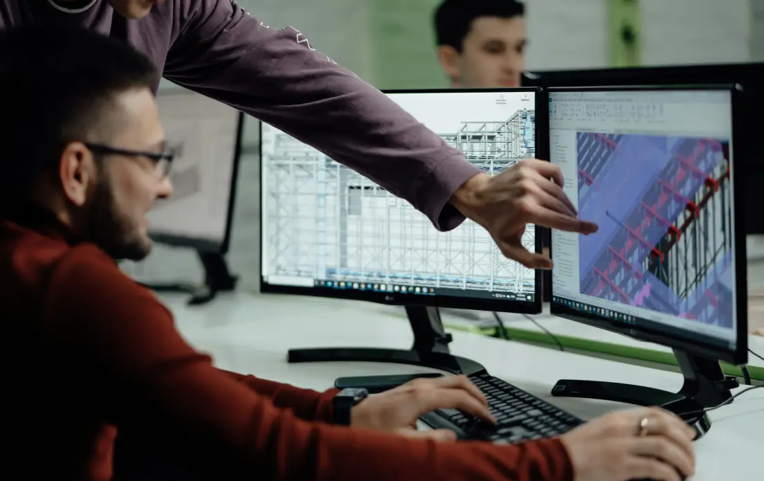 Two male engineers or designers collaborating at a workstation with dual monitors. The man in the foreground is seated, wearing glasses and a dark red sweater, with his hands on the keyboard. A colleague standing behind him in a dark purple shirt is pointing at one of the screens. The monitors display detailed 3D structural engineering or architectural models, showing what appears to be steel framework and building components in CAD software. The setting is a modern office environment